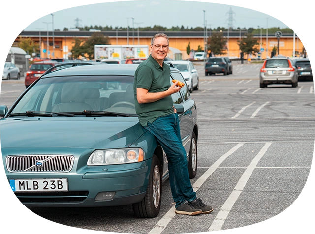Äldre glad man står lutad mot motorhuven på en volvo med en varmkorv i handen