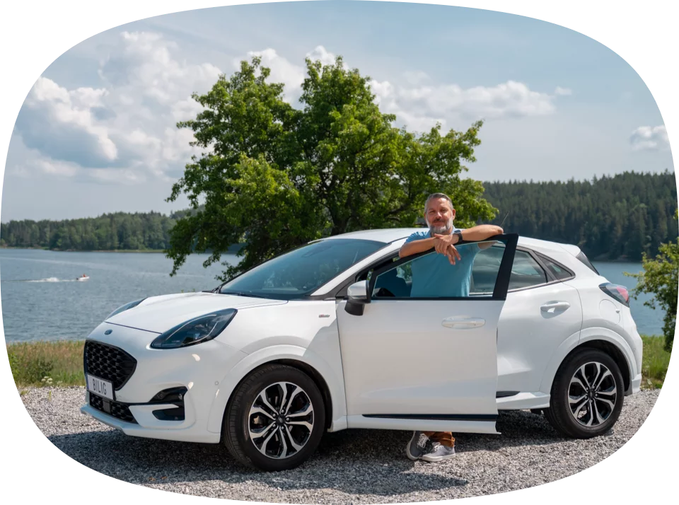 En man står bredvid en vit bil, med dörren öppen, vid en sjö. Han lutar sig mot dörren och ler mot kameran. )